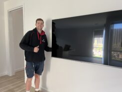 Electrician smiling and giving thumbs up beside a newly wall-mounted flat-screen TV installed in a modern home.